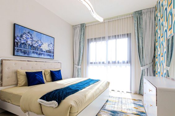 Bright and stylish bedroom in Eleganz by Danube, featuring a plush king-size bed with soft beige and blue accents, artistic decor, and large windows flooding the room with natural light.